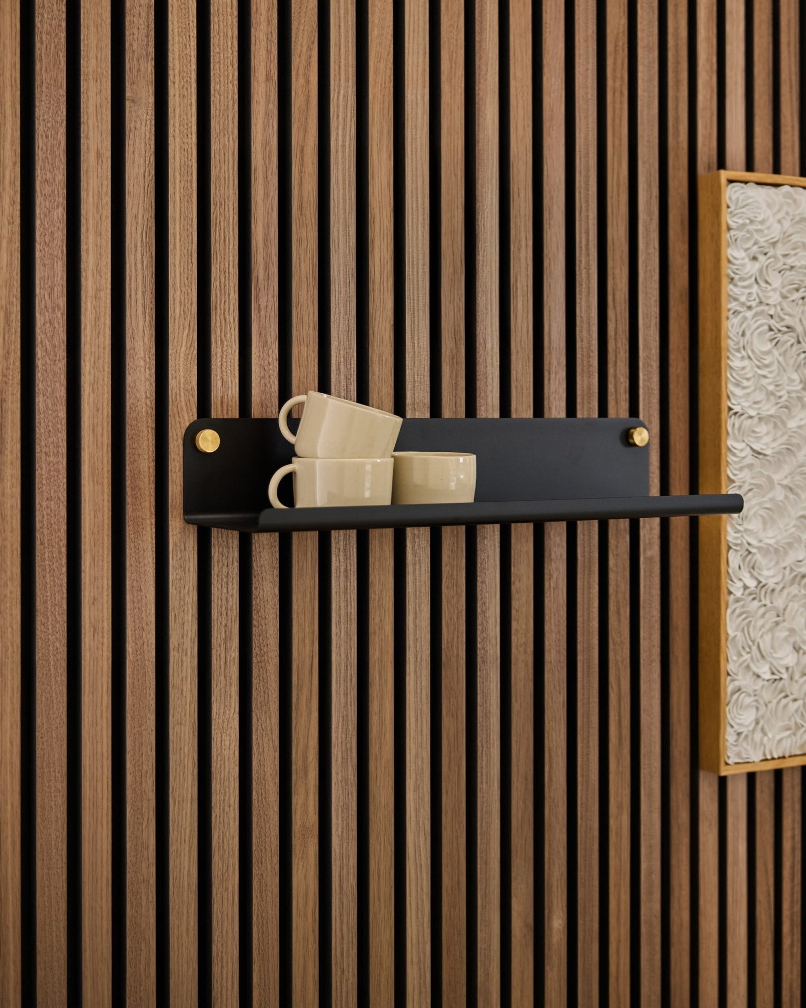 A black Norr Wide Shelf by WoodUpp holds three beige mugs, mounted on an Akupanel acoustic panel next to framed textured white artwork.